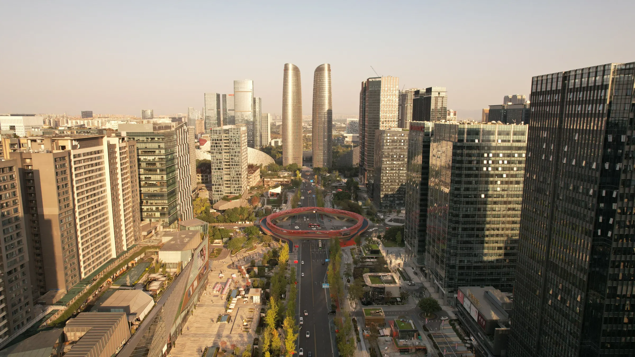 Sunlit boulevard between glass high-rises with a red ring pedestrian bridge